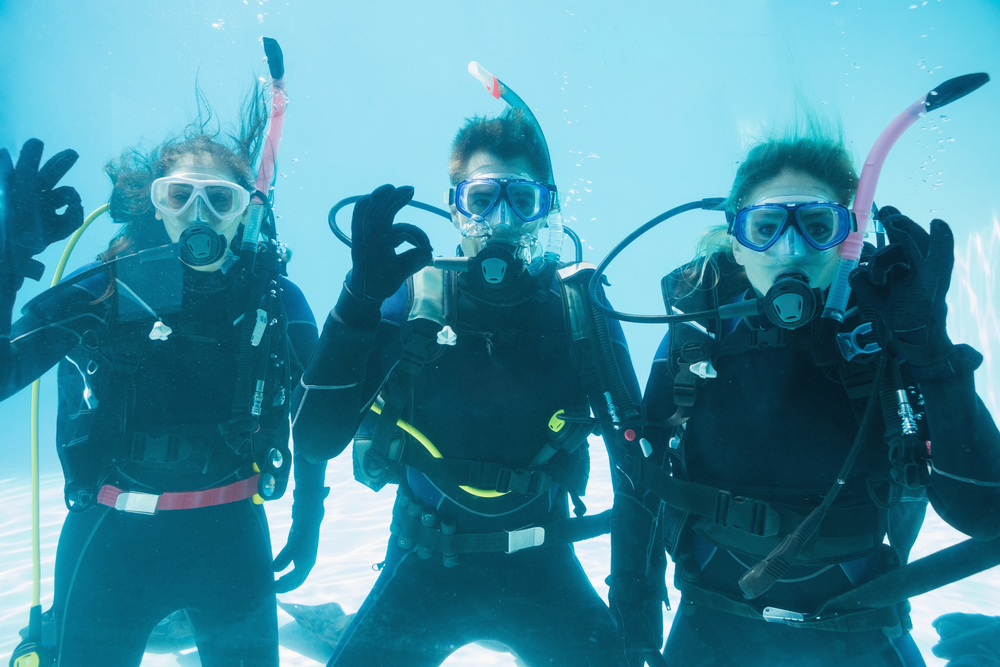Friends on scuba training submerged in swimming pool looking to camera on their holidays